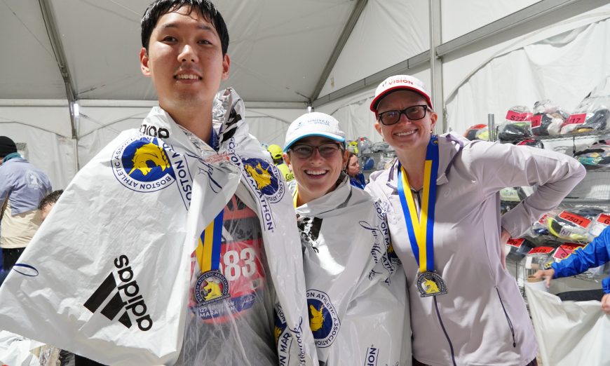 David Han in a white post-marathon wrap with two other runners inside a tent after the Boston Marathon
